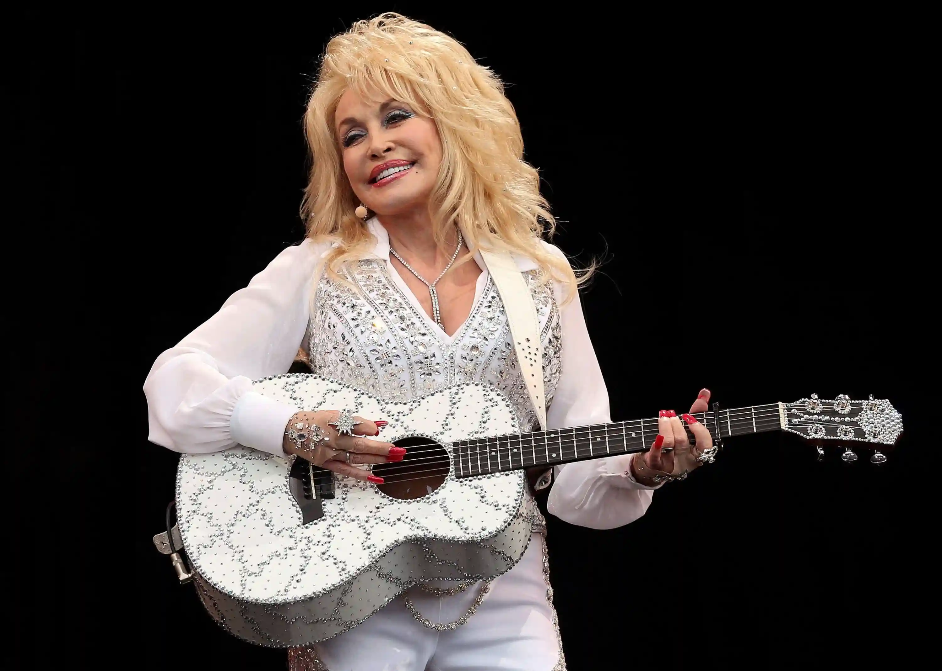 Country music legend Dolly Parton smiling warmly, holding an acoustic guitar.