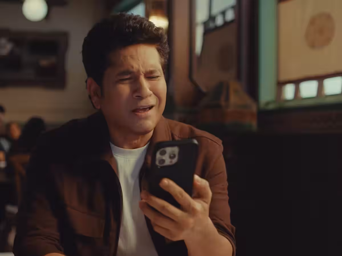 Sachin Tendulkar holding a smartphone with the ChatGPT interface on the screen, representing his new multi-year partnership with OpenAI to promote everyday AI in India.